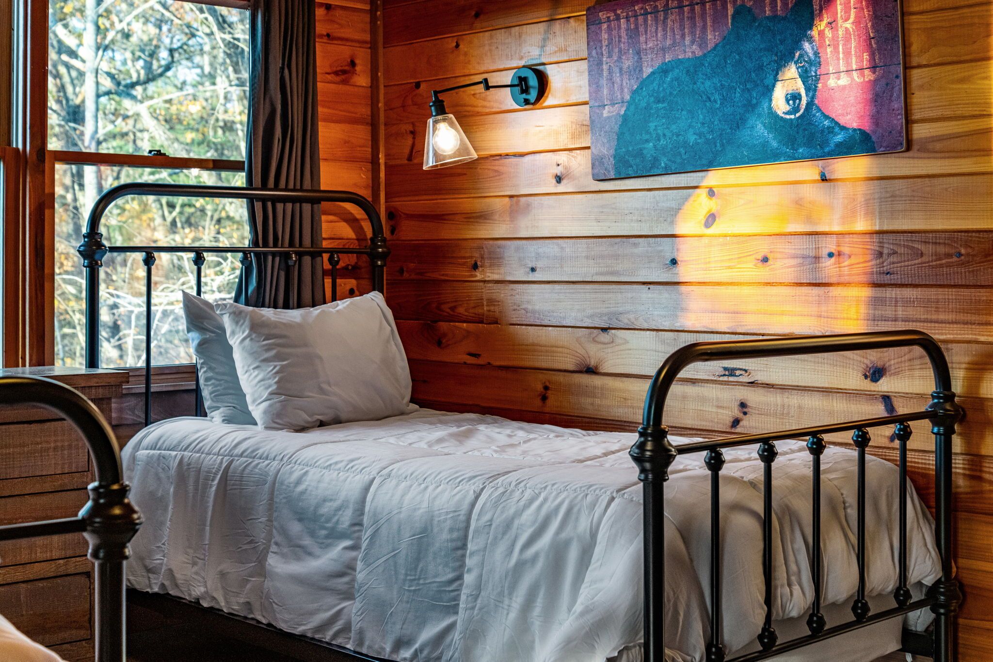 A wooden cabin-style room with a metal bed frame, a single bed with white bedding, a wall-mounted lamp, and a bear painting.