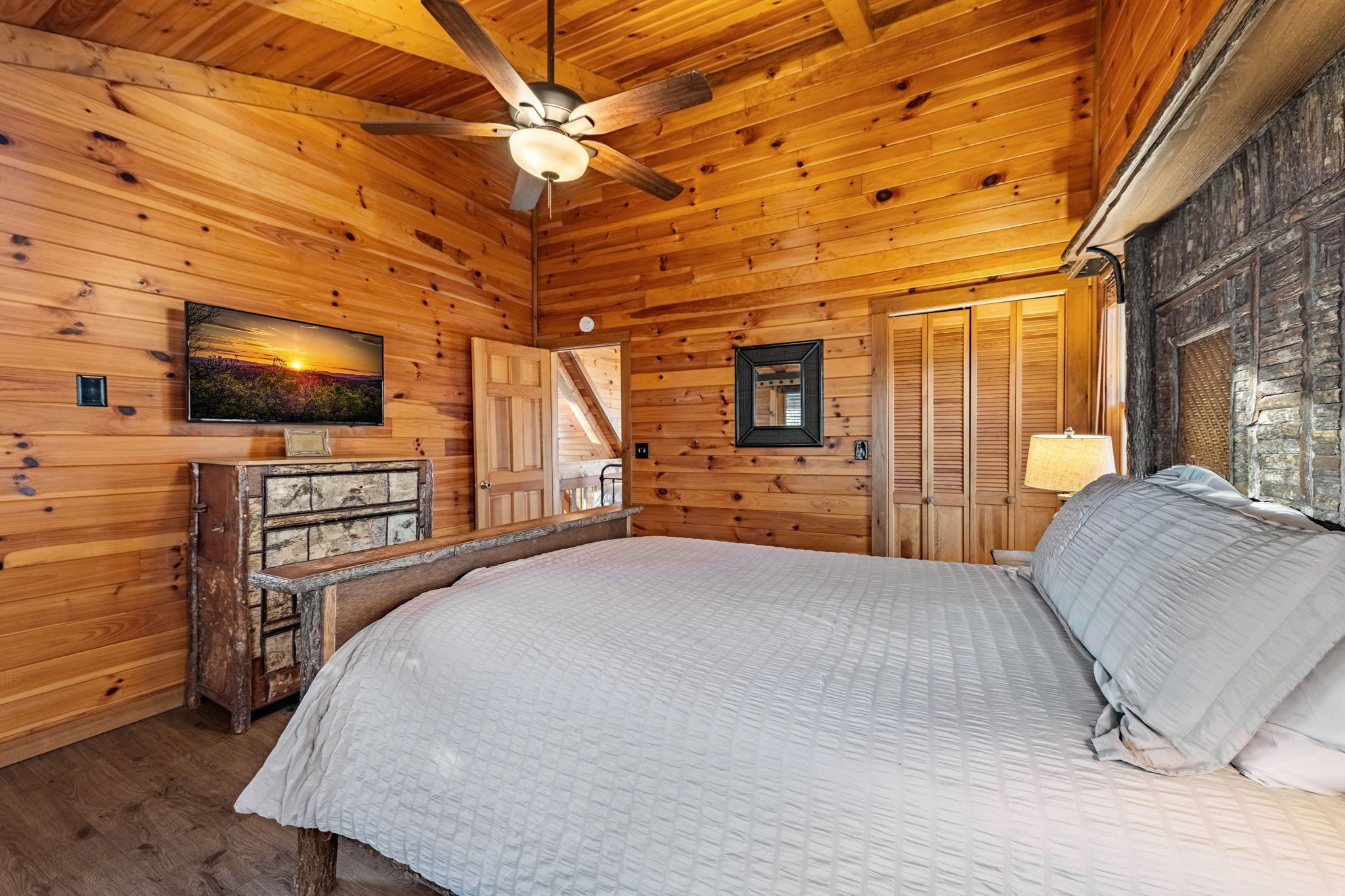 A wooden cabin room with a bed, a ceiling fan, a TV, a dresser, and a window.