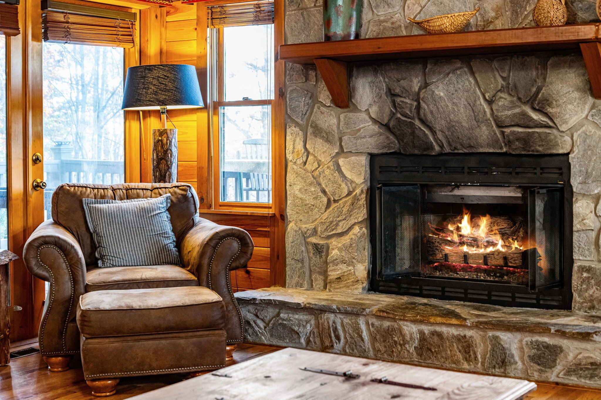 A stone fireplace with a fire, a leather armchair with a striped pillow, and a lamp.