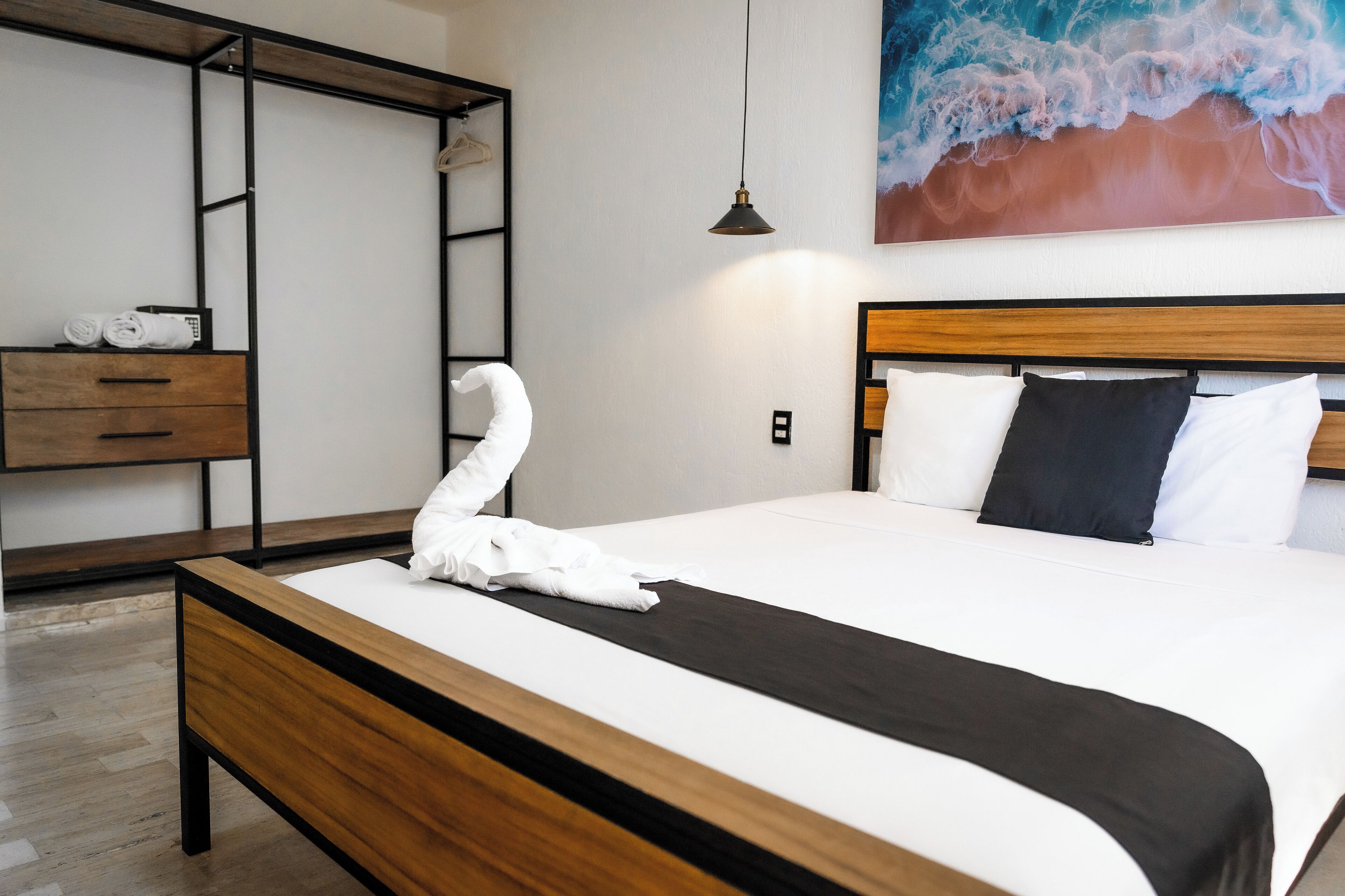 Una cama bien hecha con una toalla, una mesita de noche de madera, un banco, un estante en la pared y un cuadro de una ola.
