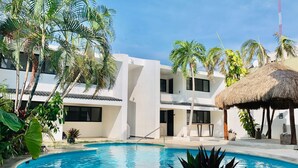 Un área de piscina con una estructura de techo de paja, rodeada de palmeras y un edificio blanco con varias ventanas.