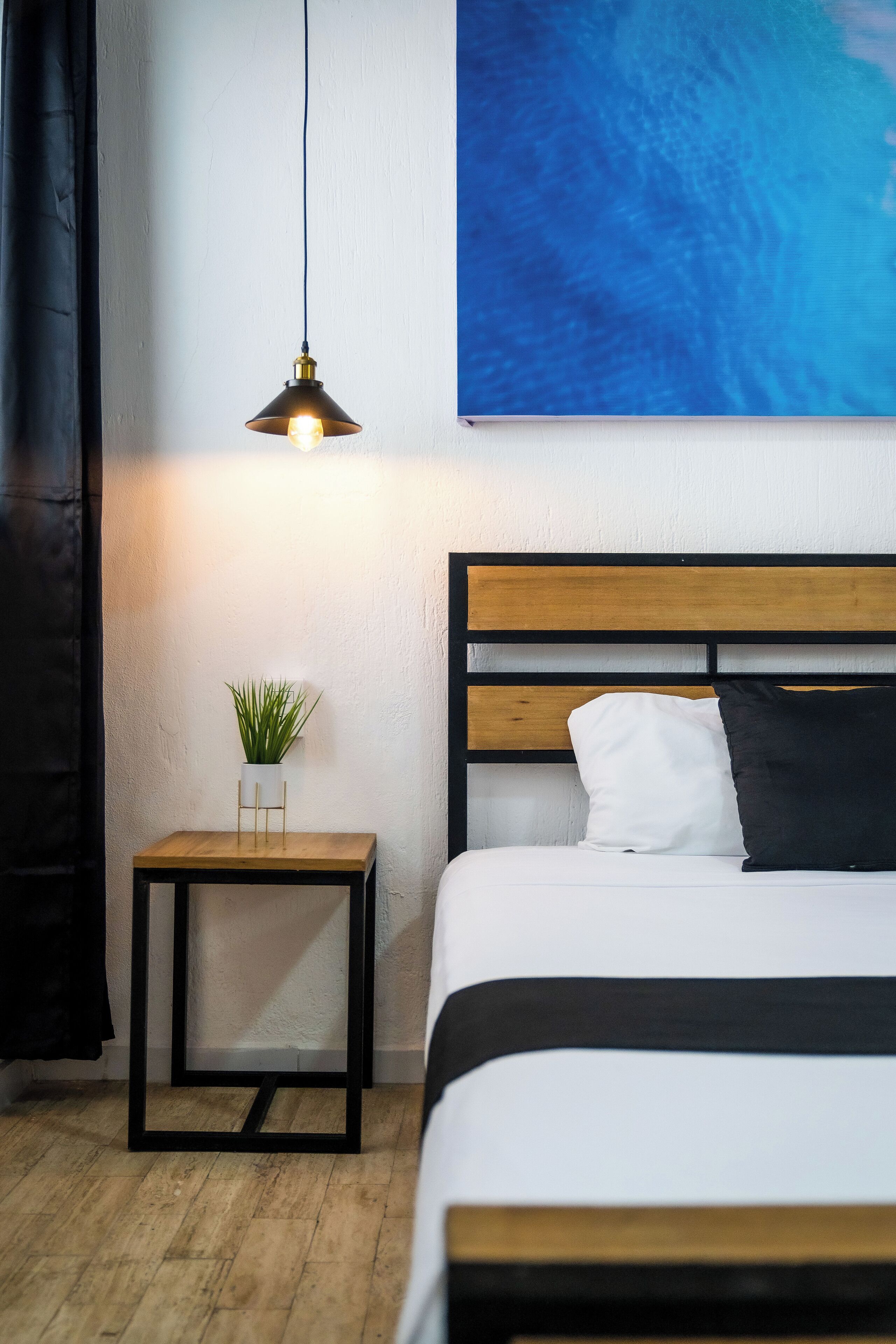 A neatly arranged bedroom with a bed, a bedside table with a plant, a hanging lamp, and a large abstract painting on the wall.
