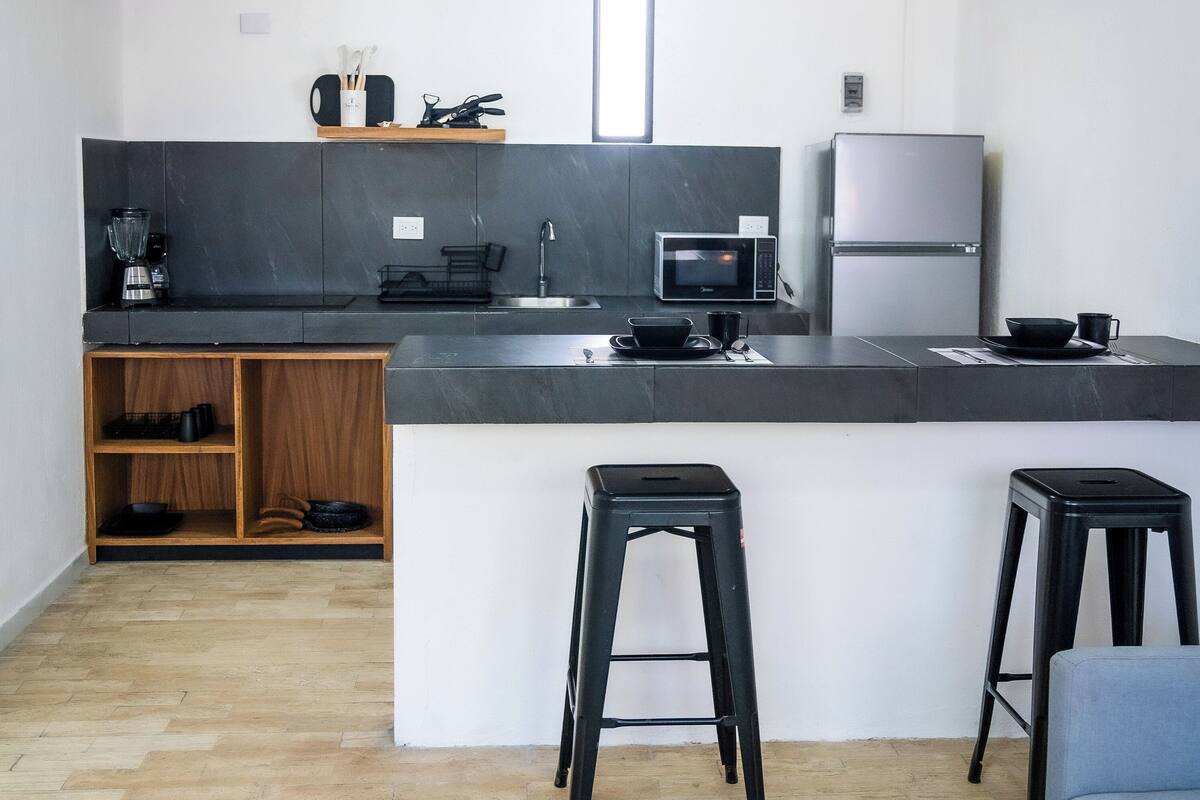 A modern kitchen with a central island, black stools, a microwave, a refrigerator, and a small sink.