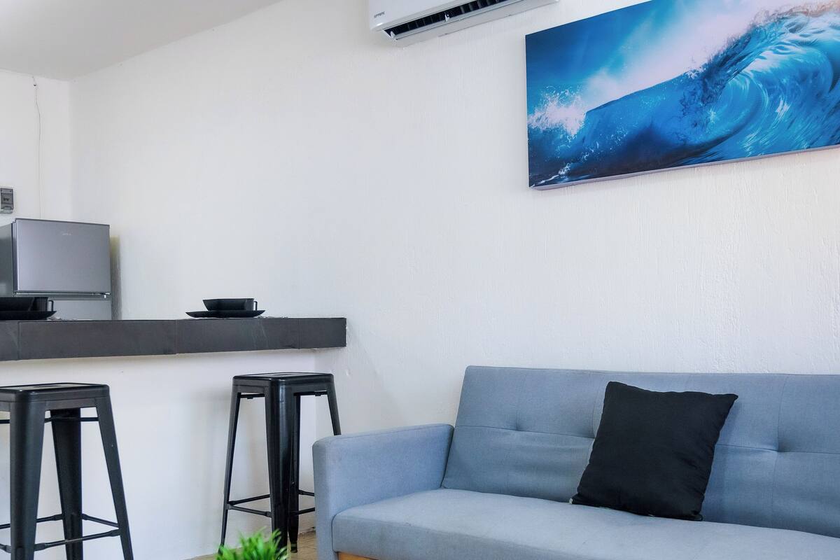A modern living room with a grey sofa, a wooden coffee table, black bar stools, a flat-screen TV, and a large wave painting on the wall.