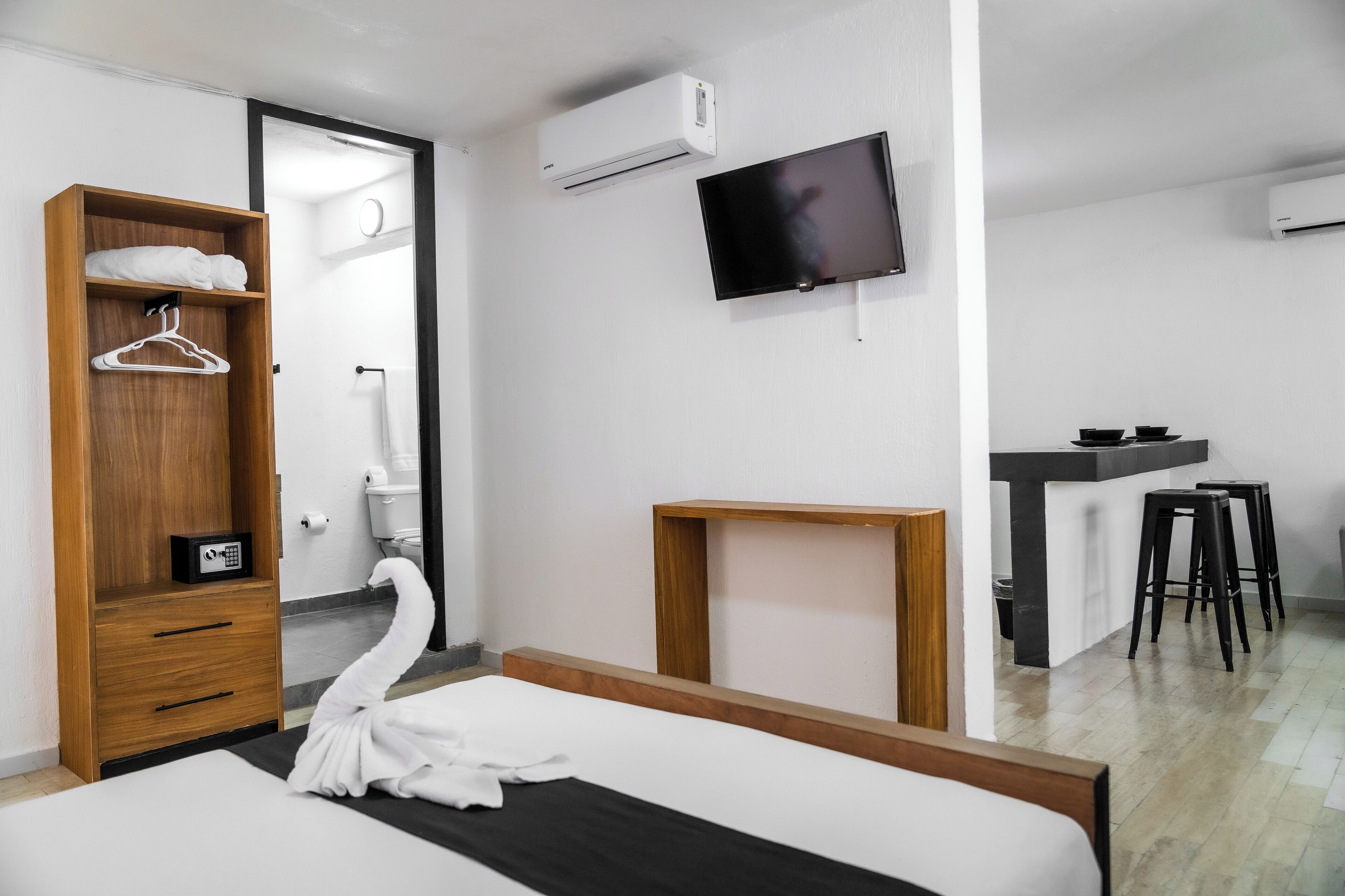 A hotel room with a bed, a wooden wardrobe, a TV, a small wooden table, and a kitchenette area.