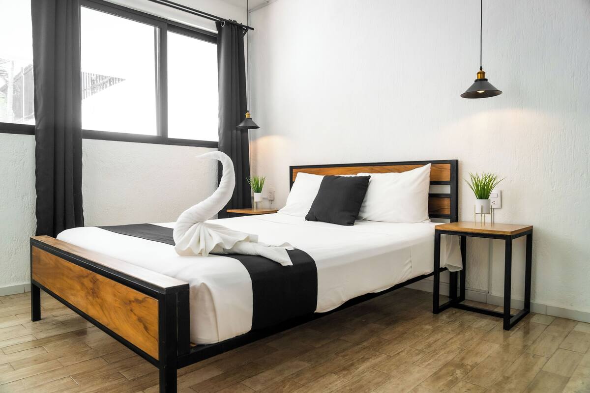 A neatly made bed with a wooden headboard, a bedside table with a lamp, and a view of the outside through a window with curtains.