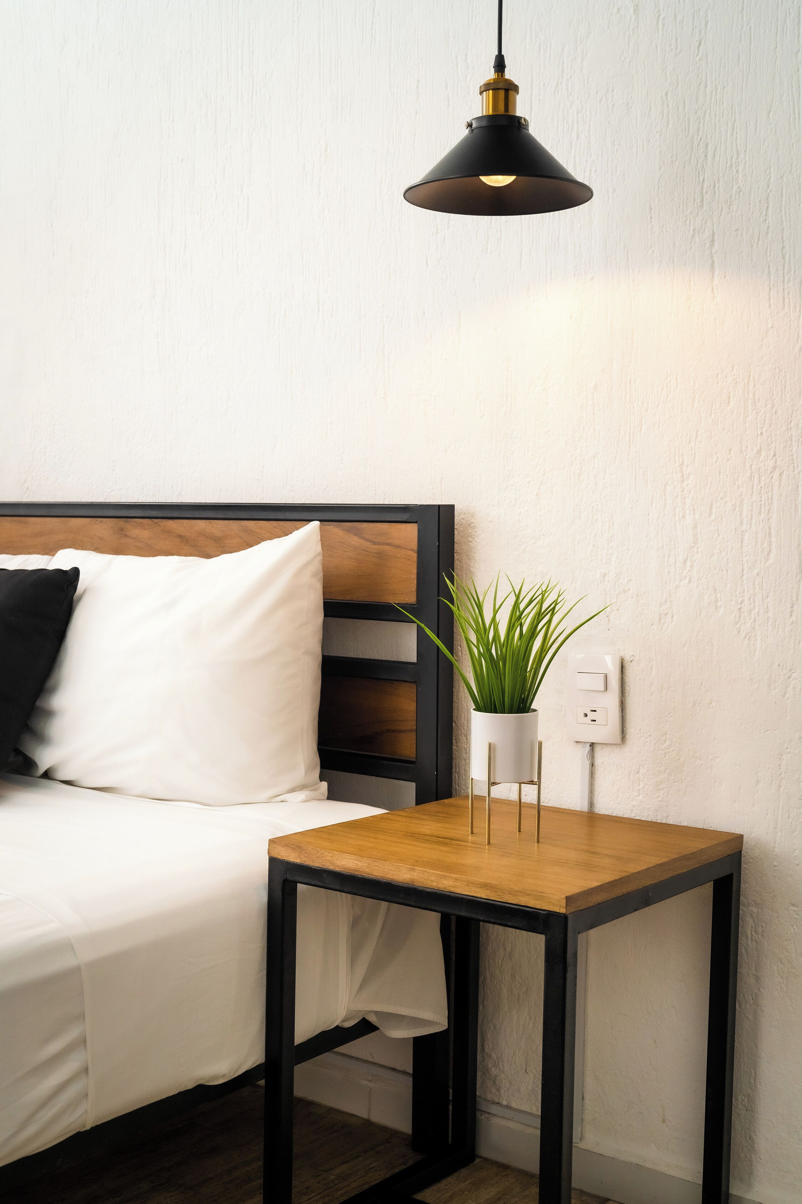 A neatly arranged room with a bed, a small wooden table with a potted plant, and a hanging light fixture.