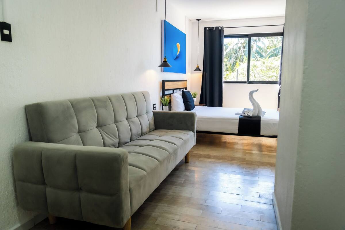 A modern room with a beige sofa, a bed with a black headboard, a blue artwork, and a window with curtains.