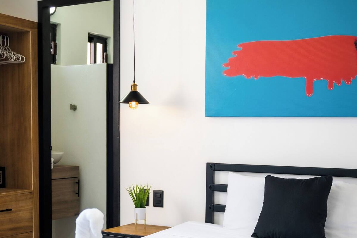 A neatly made bed with white bedding, a black headboard, and a single black pillow. A small bedside table with a lamp and a potted plant is visible. A large mirror is mounted on the wall above the bed.