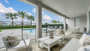 A modern outdoor patio with a pool, white furniture, and large windows offering a view of palm trees and houses. - 8BR Spectacular Villa • Games Room • Near Disney (Kissimmee)