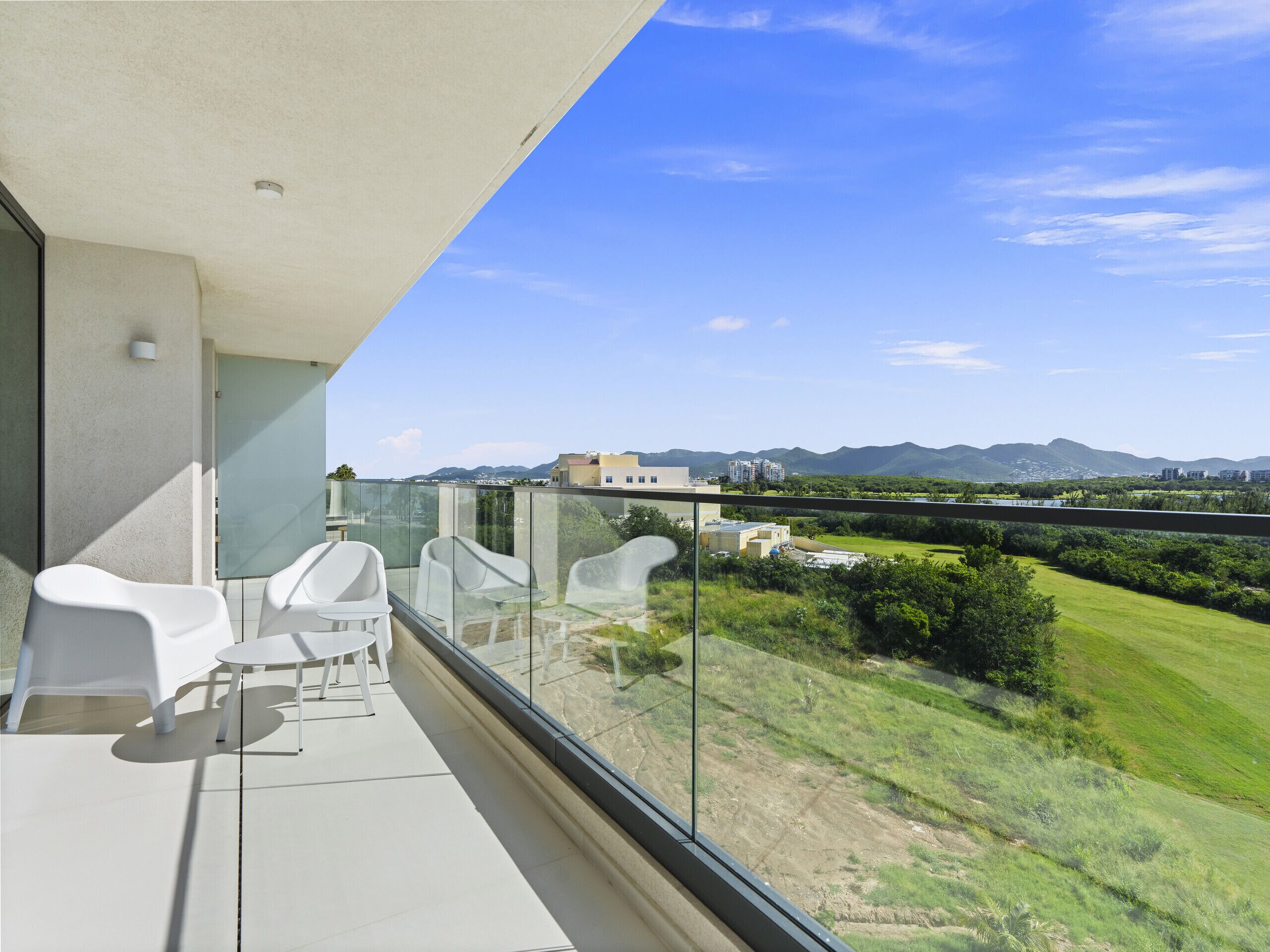 Een balkon met witte stoelen en een glazen balustrade, met uitzicht op een groen landschap en verre gebouwen.