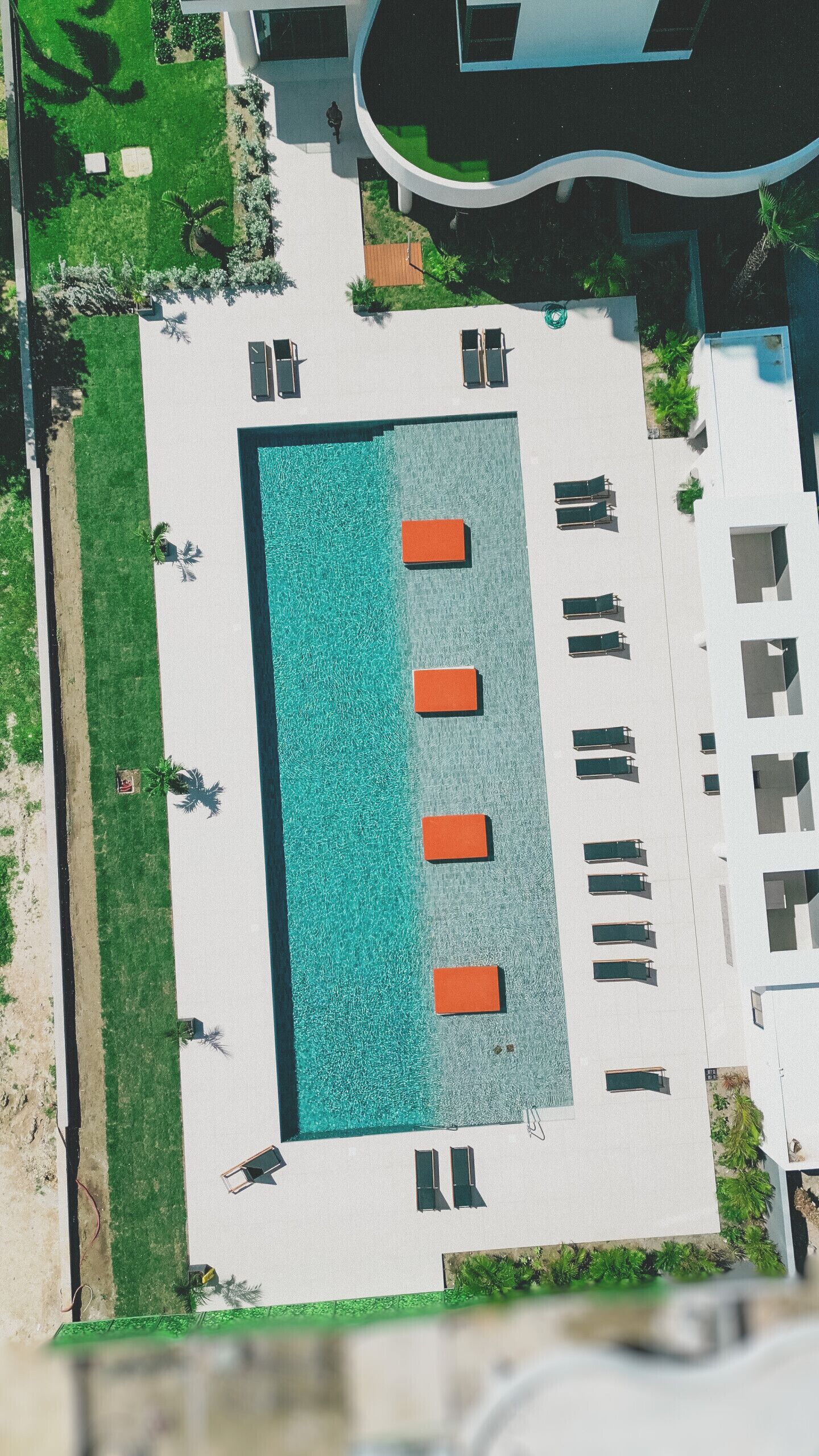 An aerial view of a swimming pool area with four sun loungers aligned along one side, surrounded by a white deck and green landscaping.