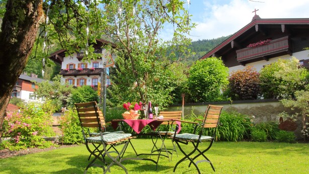 A garden with a table and chairs, surrounded by lush greenery and a building with a cross on top. - Drei-raum-ferienwohnung Winklmoos, 65qm, Dusche/wc, Extra Bad, Extra Schlafraum, Küche, Balkon (Reit im Winkl)