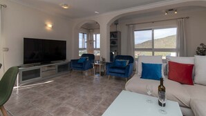 Salón con televisor, dos sillones azules, un sofá blanco con cojines rojos y azules, una mesita con una copa de vino, y vista a un paisaje montañoso a través de amplios ventanales.