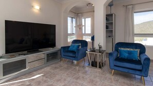 Una sala moderna con un televisor de pantalla plana grande, dos sillones azules, una mesita auxiliar y una lámpara de pie alta.