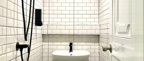 Um banheiro pequeno com azulejos brancos, pia de pedestal, chuveiro manual e sistema de ventilação visível.