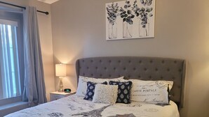 Una cama bien hecha con edredón blanco, cabecera gris y almohadones decorativos. Se ve una mesita de noche con una lámpara y un reloj.