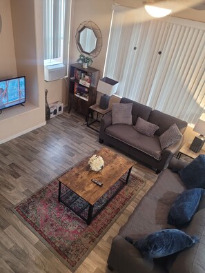 A living room with a brown sofa, a coffee table, a TV, a bookshelf, and a window with blinds.