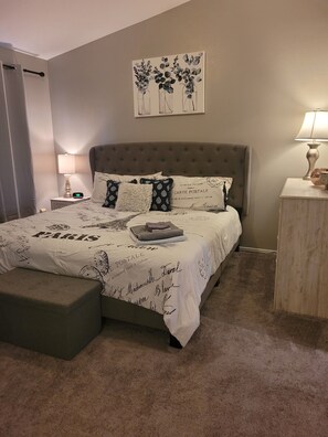Een net opgemaakt bed met een beddegoed met Parijse thema’s, een nachtkastje met een lamp en een bloemrijk kunstwerk aan de muur.