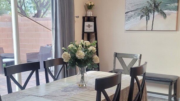 Comedor con una mesa dispuesta para cuatro, un jarrón de flores, un cuadro en la pared y vistas al exterior a través de una ventana.