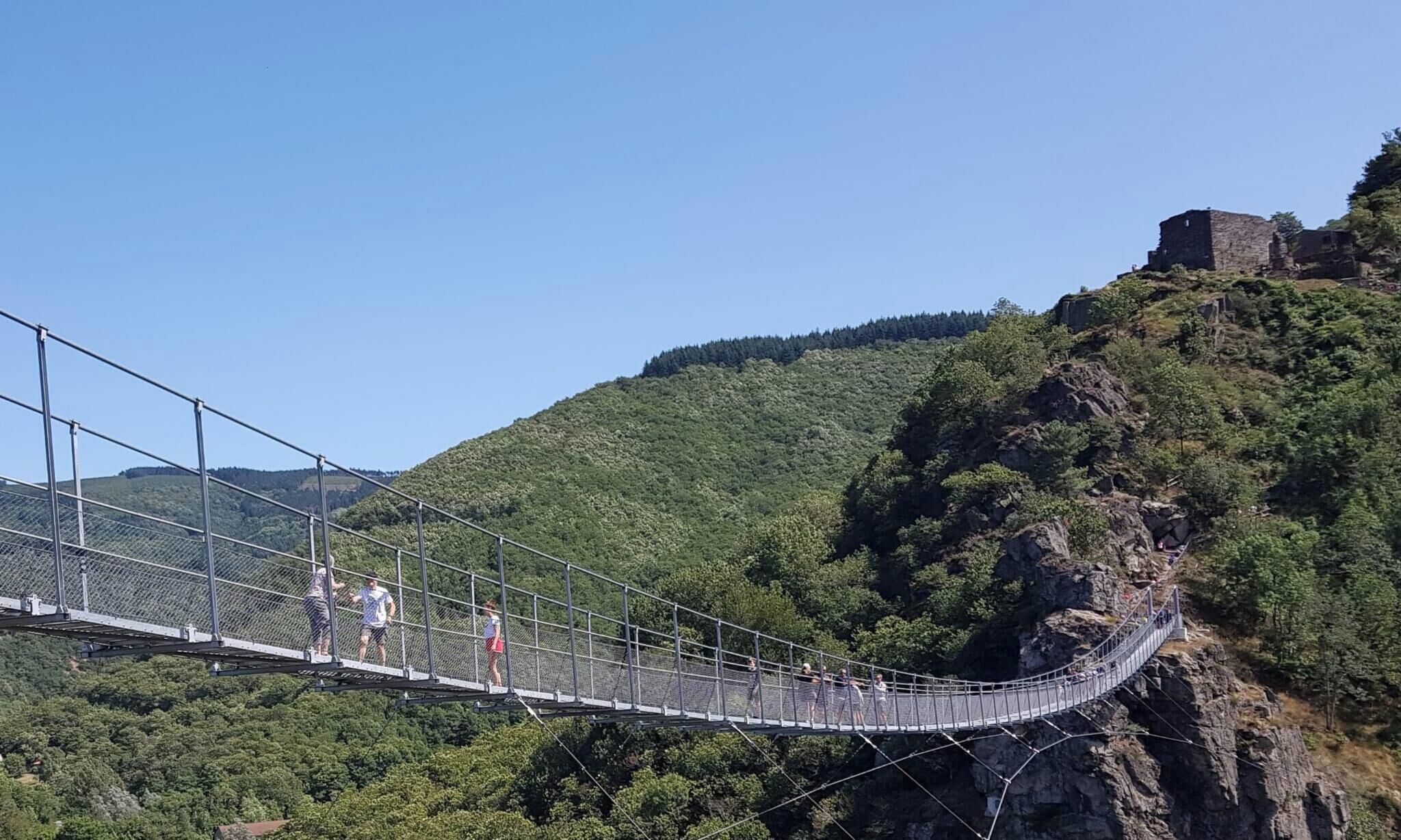 Un puente colgante con barandillas metálicas y pasarela de malla, que cruza un profundo desfiladero con una colina boscosa a un lado y una estructura de piedra histórica al otro.