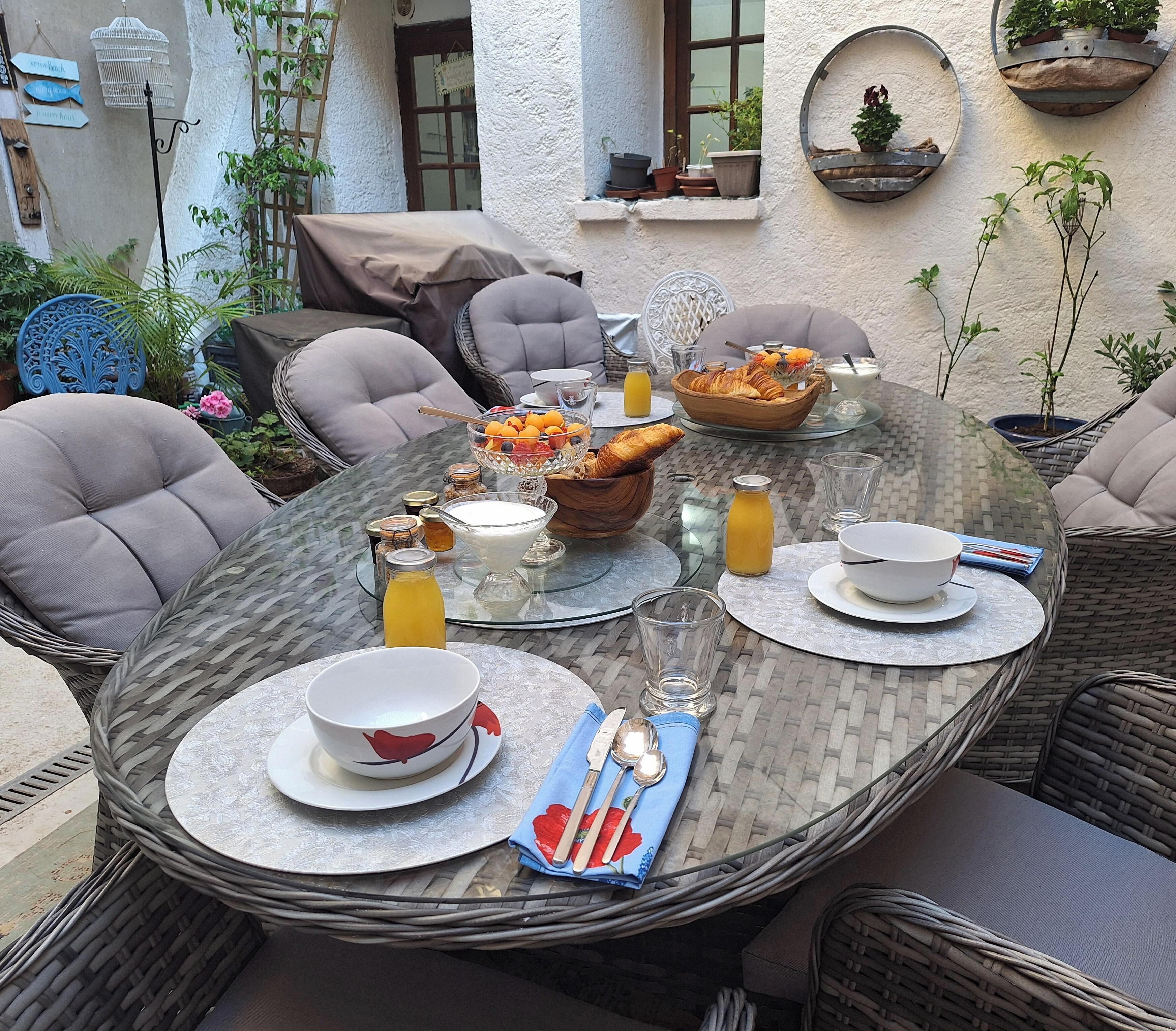 Une table ronde en verre, mise pour le petit-déjeuner, entourée de chaises en osier, dont une avec un coussin, et une petite plante en pot accrochée au mur.
