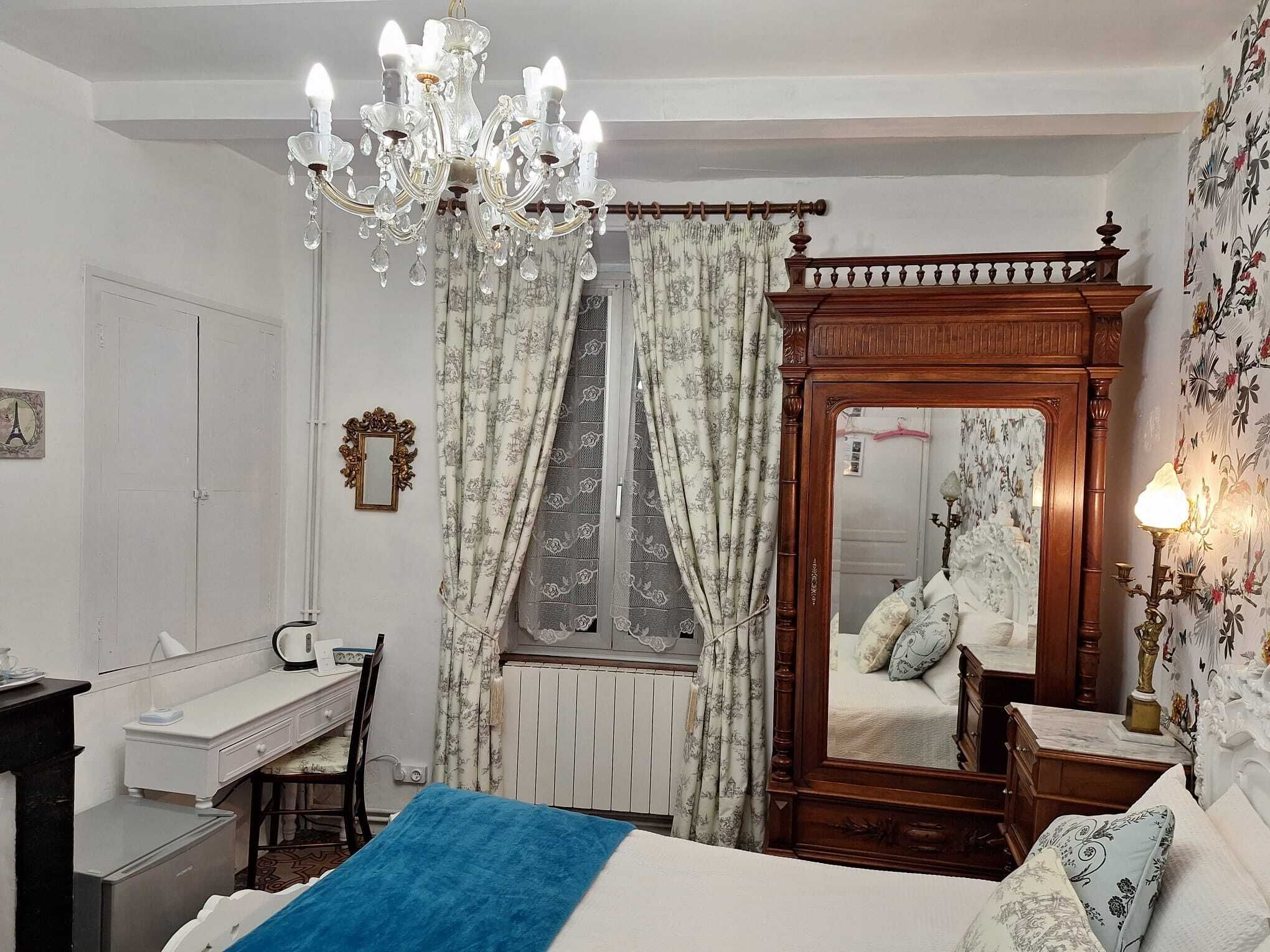 A bedroom with a large bed, a wooden wardrobe, a desk, a chair, a chandelier, and a mirror.