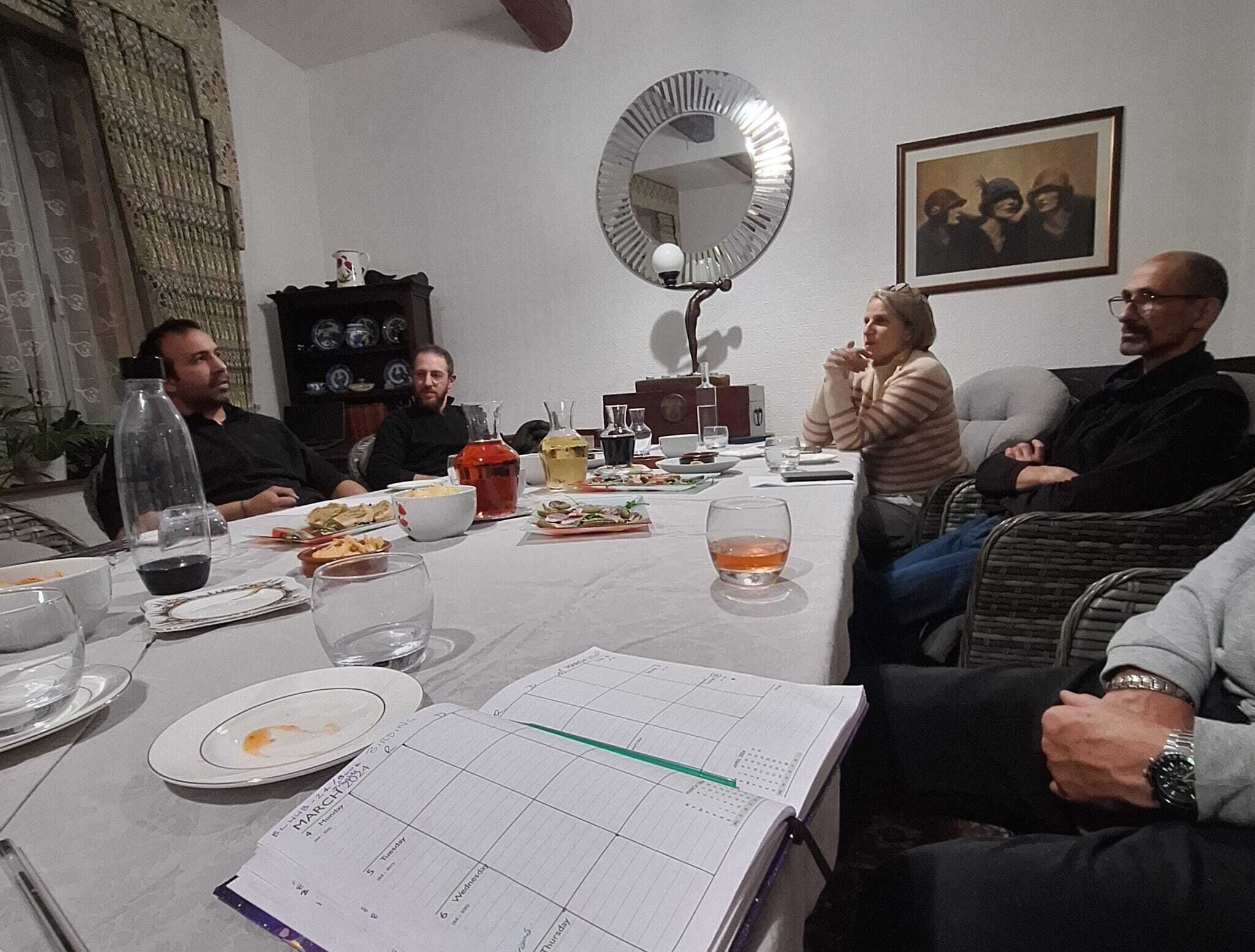 A group of people seated around a table with food and drinks, a calendar, and a mirror on the wall.