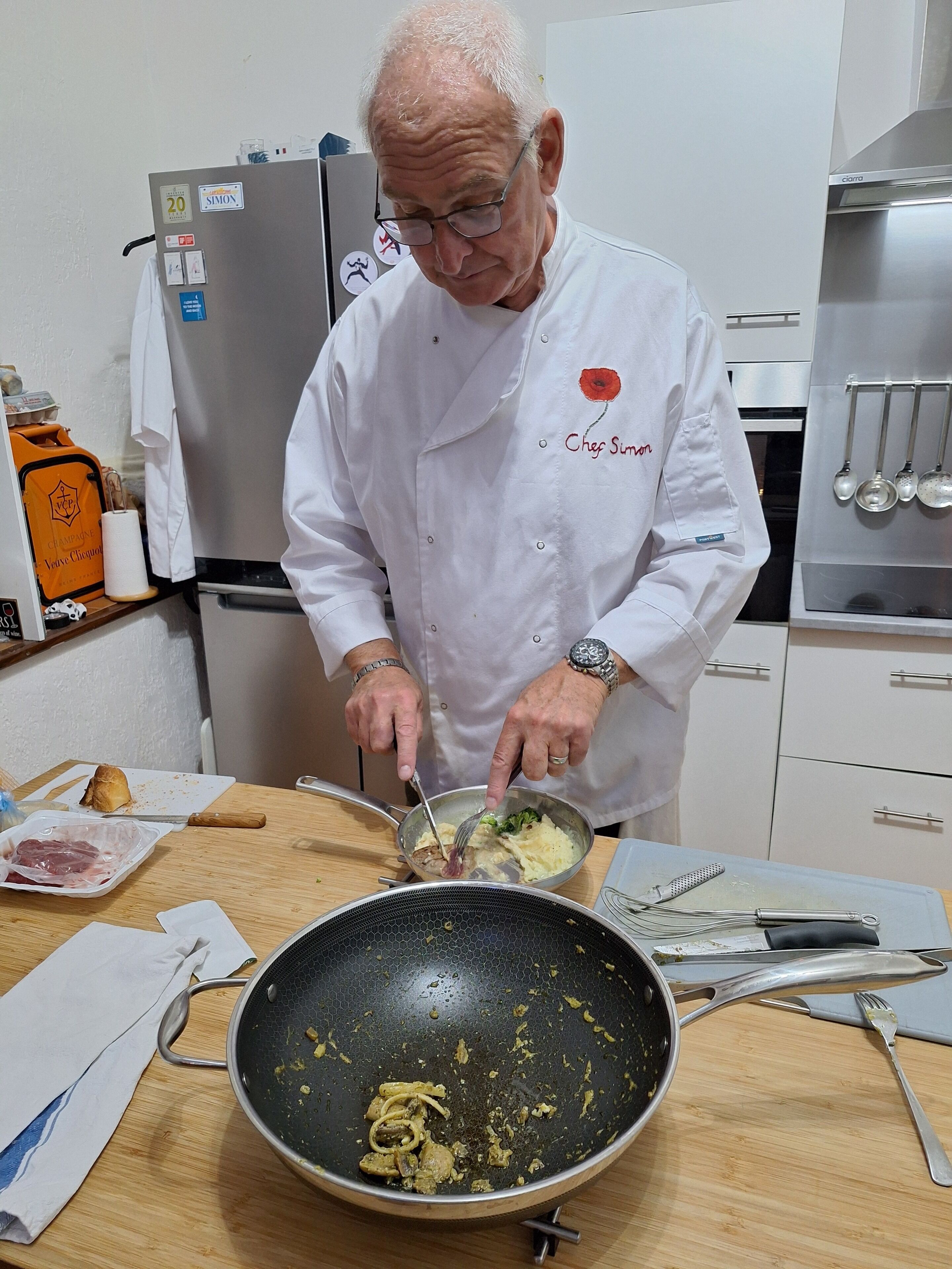 A chef in a white uniform with a red flower embroidery, preparing food in a kitchen with stainless steel appliances and utensils on the counter.