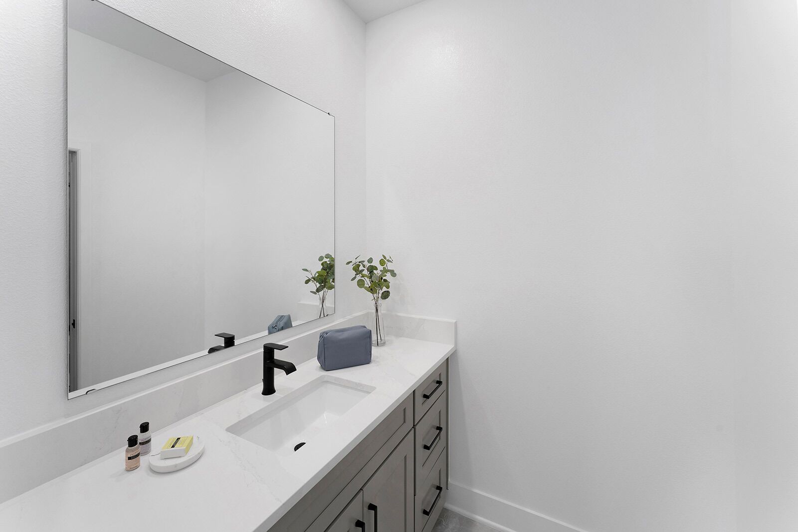A modern bathroom with a white sink, black faucet, and a large mirror. There are two small plants on the counter and a gray towel.