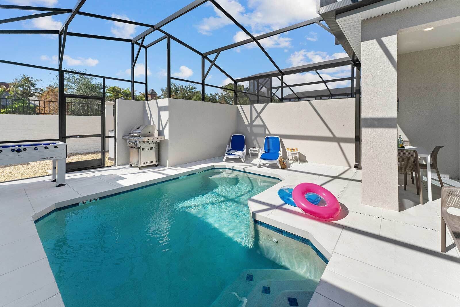 A swimming pool with a glass enclosure, lounge chairs, and a grill area.