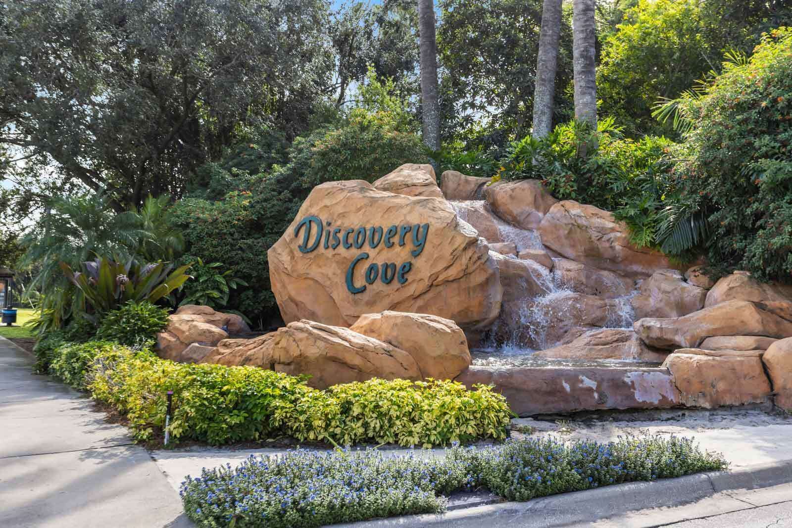 A large rock with "Discovery Cove" written on it, surrounded by lush greenery and a small waterfall.