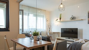 Uma sala de estar moderna com uma mesa de jantar de madeira, cadeiras brancas, um sofá, uma TV e uma bicicleta decorativa na parede.