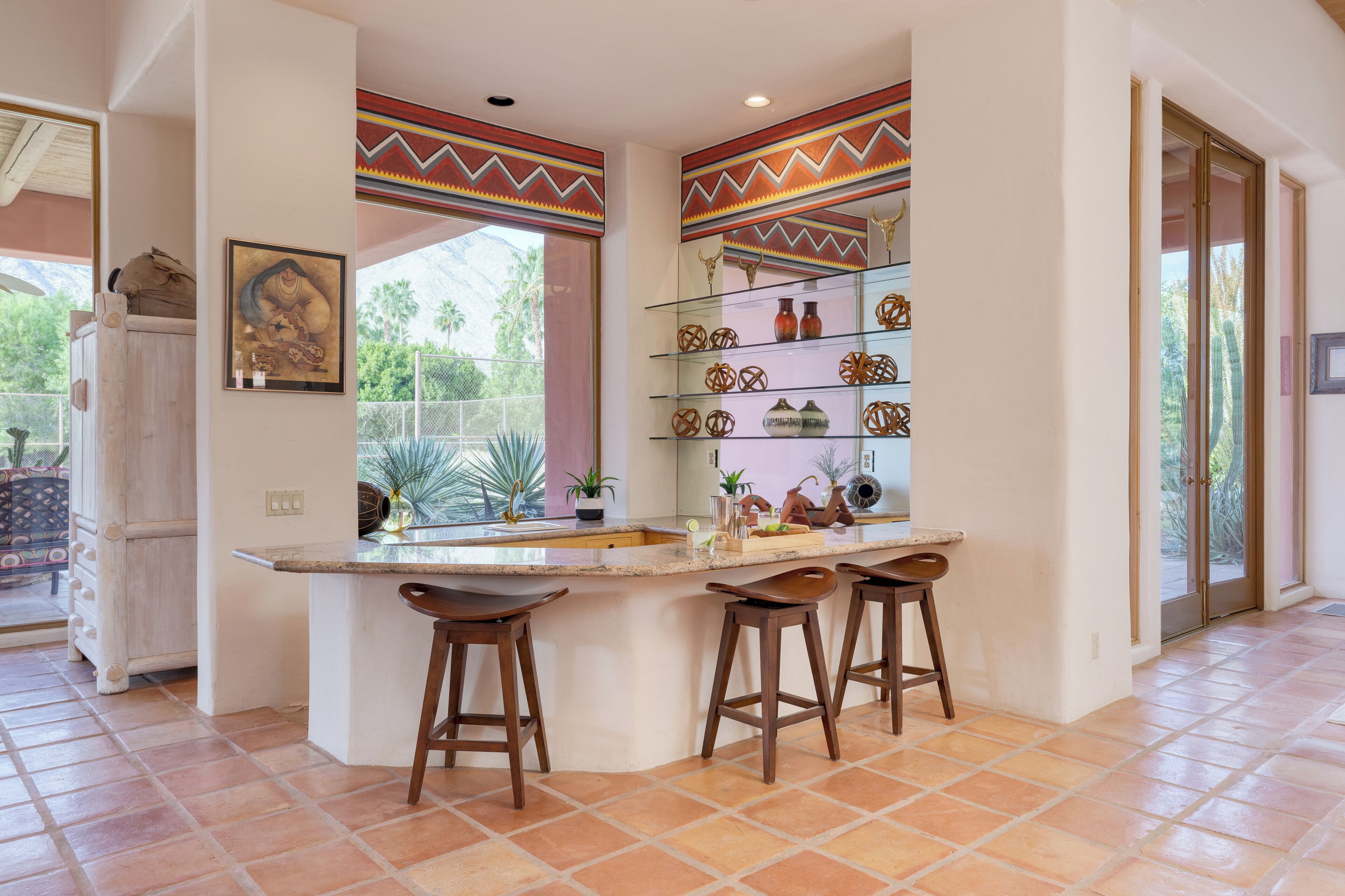 A kitchen with a central island, bar stools, a glass display shelf with decorative items, and a window with a view of greenery.