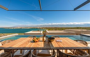Una mesa de madera con sillas de mimbre, con vista a una piscina y un paisaje con un lago y montañas.