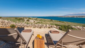 Blick auf einen Strand mit zwei Liegestühlen, einem kleinen Holztisch mit Orangensaft und einer Orange, und einer Steinmauer im Vordergrund.
