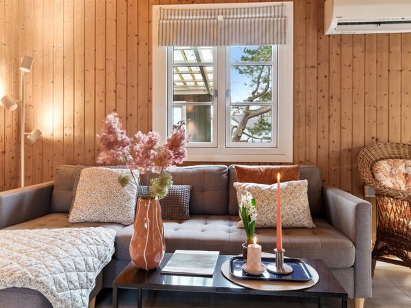 Un salon chaleureux avec un mur lambrissé en bois, un canapé gris agrémenté de coussins à motifs, un vase rempli de fleurs roses et une bougie allumée posée sur un plateau.