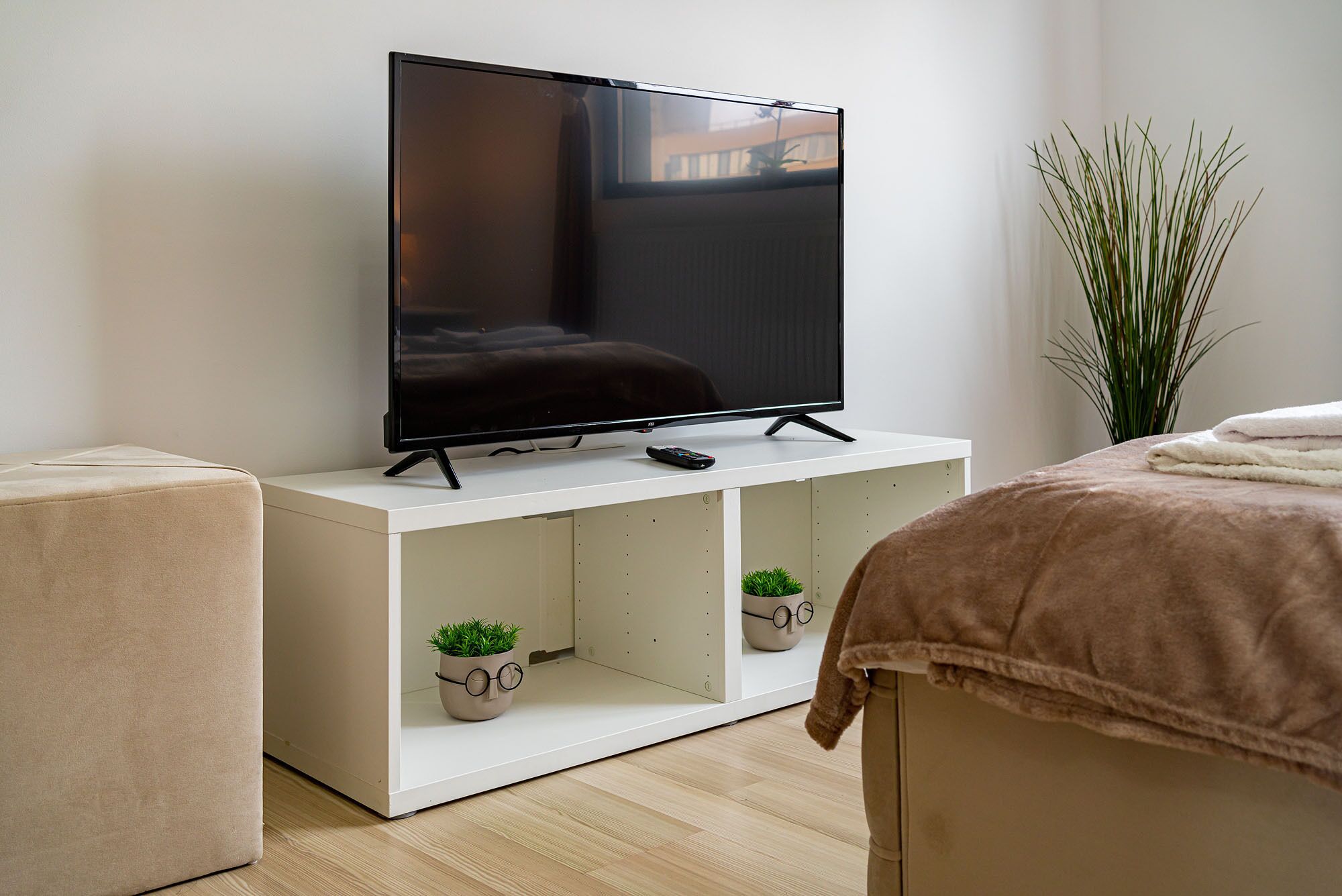Una sala de estar moderna con un televisor de pantalla plana sobre un mueble blanco, un sofá color beige, una planta pequeña en maceta y una planta verde alta junto a una cama con una manta marrón.