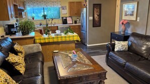 A living room with a brown leather sofa, a wooden coffee table, and a kitchen area with wooden cabinets and a refrigerator. - Clean safe double bedroom, comfy bed, keyless entry, no stairs, on bus routes (Langley Township)