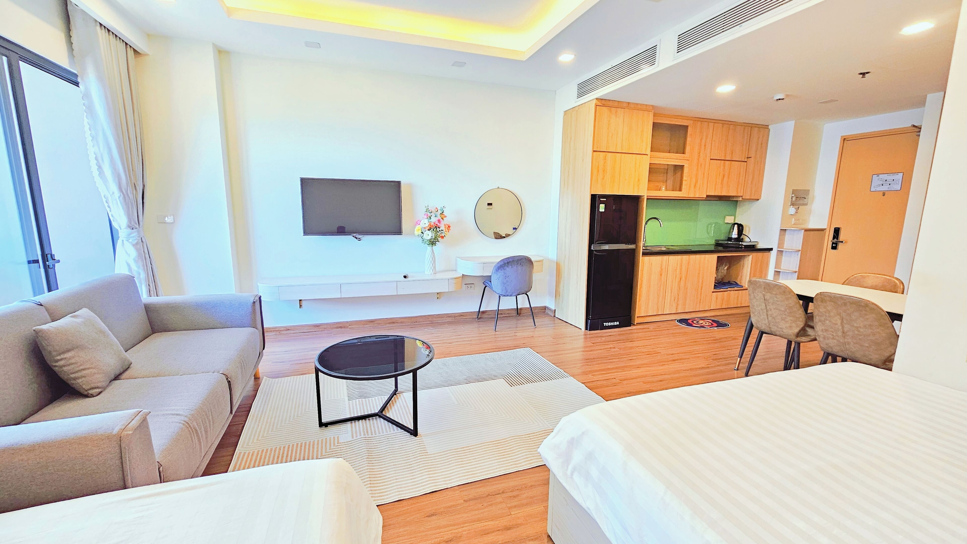 A modern hotel room with a sofa, a bed, a dining table, a kitchen area, and a flat-screen TV.