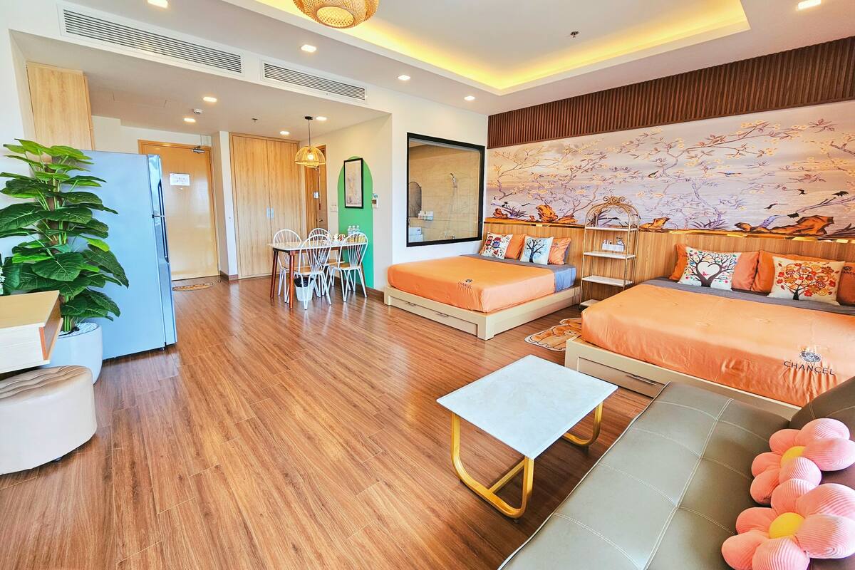 A modern hotel room with a wooden floor, a bed with orange bedding, a white sofa, a small table, and a large mirror.