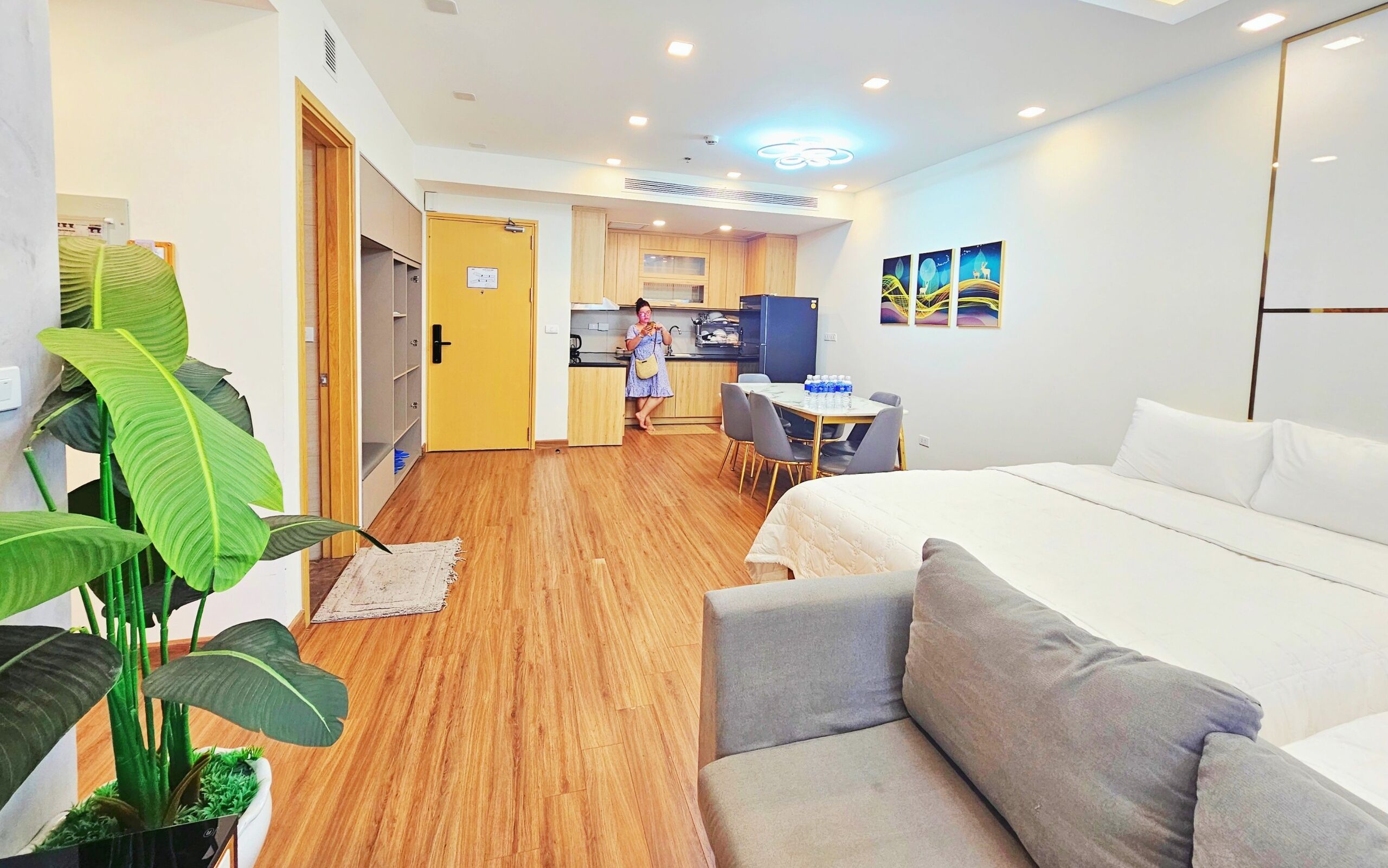A hotel room with a bed, a sofa, a dining table with chairs, a kitchen area, and a large plant.