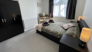 A bedroom with a bed, bedside tables, a wardrobe, a radiator, a window with curtains, and a desk with a chair.