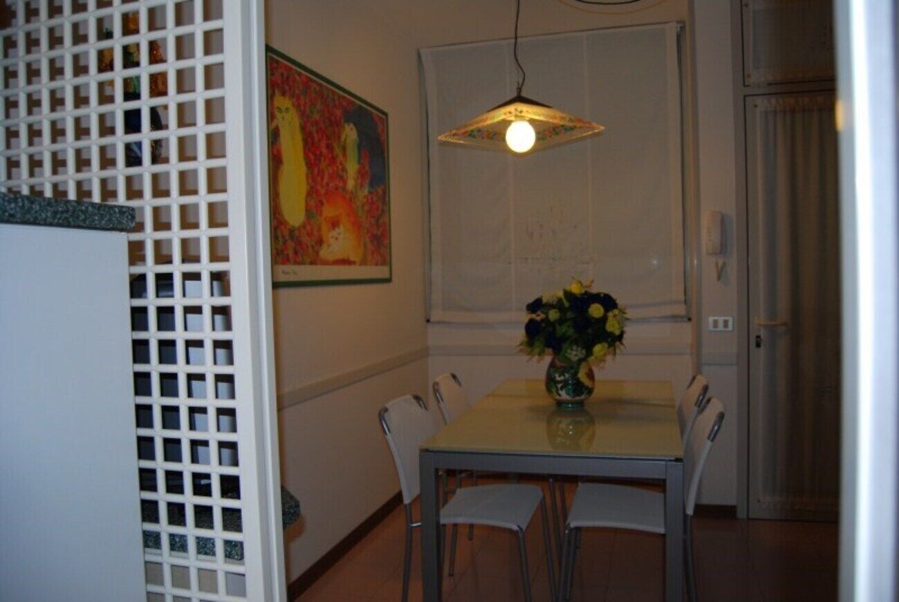 A dining area with a table, chairs, a vase with flowers, a painting on the wall, and a window with blinds.