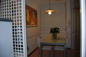 A dining area with a table, chairs, a vase with flowers, a painting on the wall, and a window with blinds. - Relaxing Apartment With Garden and Pool (San Michele al Tagliamento)