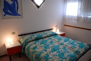 Ein Schlafzimmer mit Bett, Nachttischen, einer Wandlampe, einem Fenster mit Vorhängen und einer Deckenleuchte.
