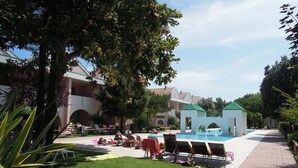Un espace piscine avec des chaises longues et un petit bâtiment, le tout entouré de verdure et bordé d’un chemin pavé.