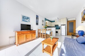Een woonkamer met een televisie, een houten kast, een eettafel en stoelen. Er is een keukenhoek met een koelkast en een deur naar een andere kamer.