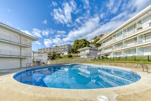 A swimming pool surrounded by modern buildings with balconies, a clear sky, and a hilly backdrop.