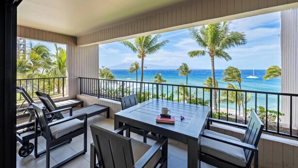 Un balcon avec vue sur l’océan, comprenant une table et des chaises, des palmiers et un voilier à l’horizon.